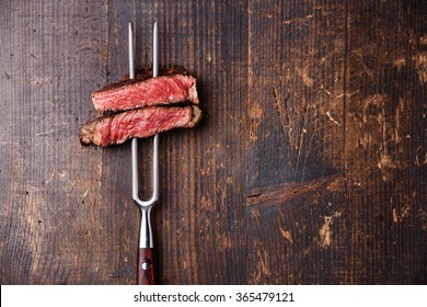 Slices Of Medium Rare Grilled Steak Ribeye On Meat Fork On Dark Wooden Background