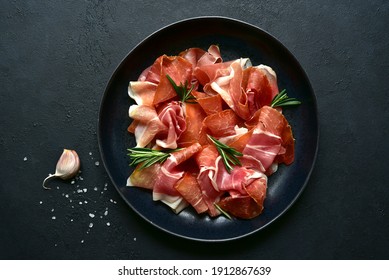 Slices of prosciutto di parma or jamon serrano (iberico)  on a black plate on a dark slate, stone or concrete background. Top view with copy space. - Shutterstock ID 1912867639