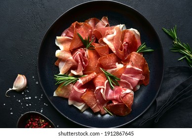 Slices of prosciutto di parma or jamon serrano (iberico)  on a black plate on a dark slate, stone or concrete background. Top view with copy space.