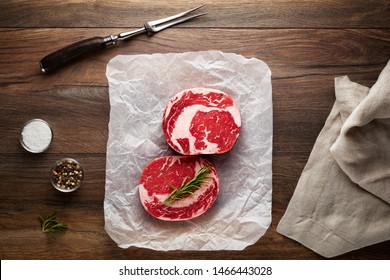 Slices Of Raw Rib Eye Steak On White Cooking Paper And Wooden Table. Decorated With Salt, Pepper, Meat Fork And Napkin. Overhead View.