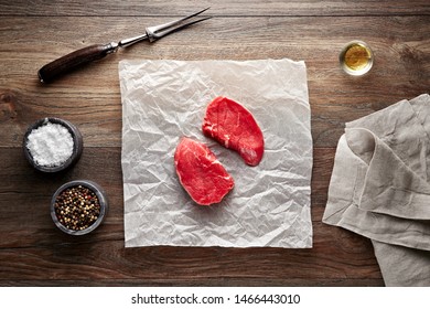 Slices Of Raw Tenderloin Steak On White Cooking Paper And Wooden Table. Decorated With Salt, Pepper, Meat Fork And Napkin. Overhead View.