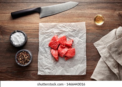 Slices Of Raw Veal Goulash Meat On White Cooking Paper And Wooden Table. Decorated With Salt, Pepper, Meat Fork And Napkin. Overhead View.