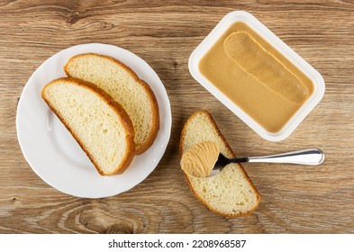 Slices Of Wheat Bread In White Plate, Teaspoon With Peanut Butter On Slice Of Bread, Plastic Box With Sweet Peanut Paste On Wooden Table. Top View