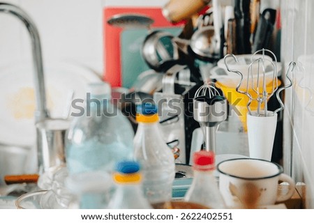 Slightly blurred view of a cluttered kitchen countertop featuring various utensils, bottles, a mug, and kitchen appliances like a blender. Represents daily kitchen life and routine.