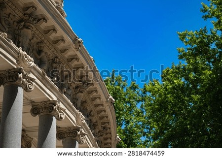 A slightly wider angle of the same neoclassical colonnade reveals more of the sweeping curved stone structure. Multiple Corinthian columns with richly carved capitals support the ornate entablature while bright summer light picks out the sculptural detail against the vivid blue sky.