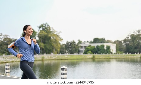 Slim and active Asian woman in sportswear listening to music while running or jogging along the lake in the beautiful nature park in the morning. - Shutterstock ID 2244345935