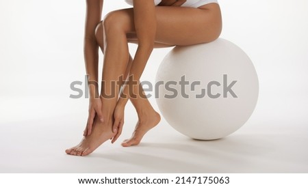 Slim African American beauty model touches her feets and legs sitting on white ball against white background | Foot care commercial concept