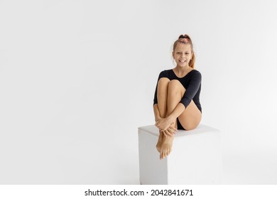 slim artistic teenager girl in black leotard trains and having fun on white cube on white background in rhythmic gymnastic exercise, children's professional sports