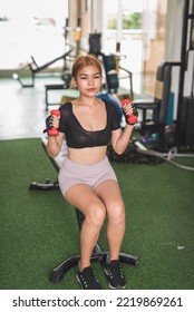 A Slim  Asian Woman Does Seated Dumbbell Hammer Curls On A Flat Bench At The Gym. Wearing A Black Crop Top And Bike Shorts.