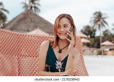 A Slim Asian Woman With Dyed Hair Calling A Friend On Her Phone While Enjoying Her Vacation At A Posh Tropical Beach Resort.