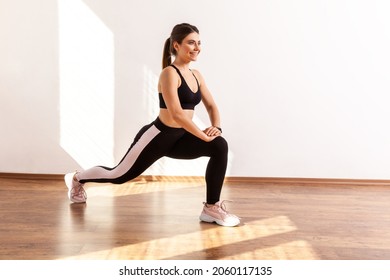 Slim Athletic Beautiful Female Stretching Legs Before Workout, Lower Body Exercise, Wearing Black Sports Top And Tights. Full Length Studio Shot Illuminated By Sunlight From Window.