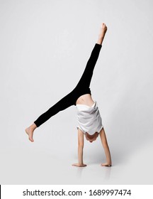 Slim Athletic Blonde Kid Girl Is Doing Gymnastic Acrobatics Exercise At Home In Studio, Handstand And Stretching With Her Legs Up On White Background