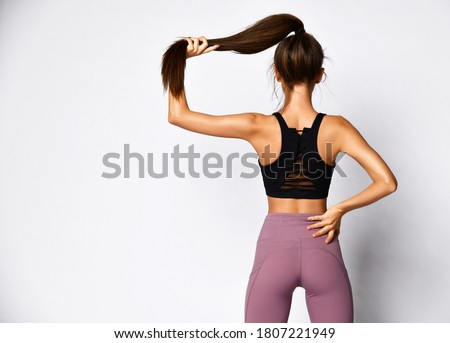 Slim athletic brunette woman in sportswear pants and sports bra top stands back to us holding ponytail in hand on gray  background