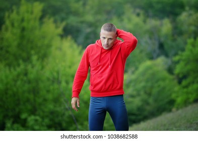 A  slim  athletic runner in red jacket with a hood and black sport leggins rest after jogging on a colorful green forest background.