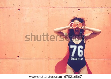 Slim attractive girl with curly hair motivating other eat healthy food fruit using grapefruit while standing on pink wall background with copy space area for your advertising messages. Summer mood