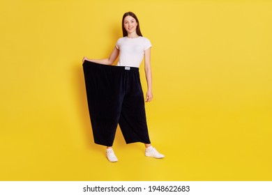 Slim Attractive Girl With Dark Hair Wearing White Caucasian T Shirt And Too Big Black Pants, Female Lost Weight And Being Proud Of It, Looks Smiling At Camera, Isolated Over Yellow Background.