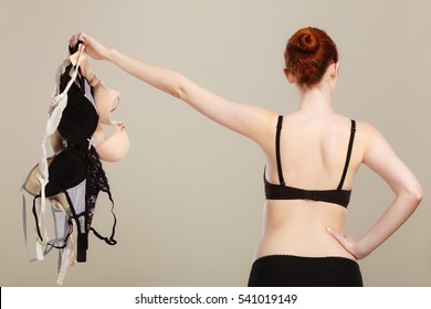 Slim attractive red hair woman wearing black underwear holding many bras in hand, choosing which bra to wear, rear view