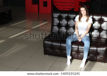Slim and beautiful young blonde posing in the studio on a black leather sofa. Pretty woman in tight blue jeans.