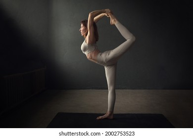 A Slim Beautifully Lighted Young  Woman Is Doing Natarajasana (Lord Shiva’s Yoga Pose) In A Dark Room. Image With Selective Focus And Toning