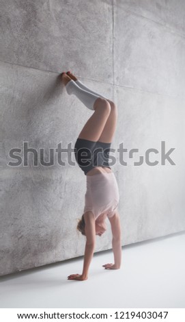 Slim blond woman in shorts doing handstand with legs on gray wall in daylight