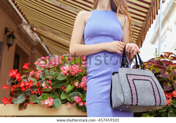 Slim blonde in pale violet sheath dress with gray\
felt bag near the blooming flowers in the summer terrace of\
cafeteria. Young stylish woman posing on the street. Pretty woman\
with long hair.