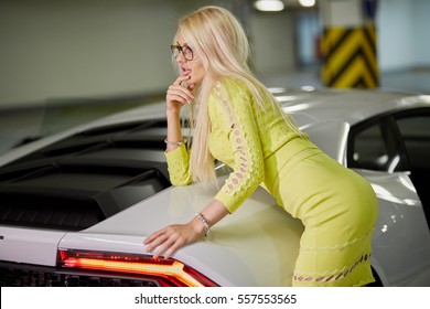 Slim blonde woman in yellow dress stands leaning on trunk of modern white car at underground parking.