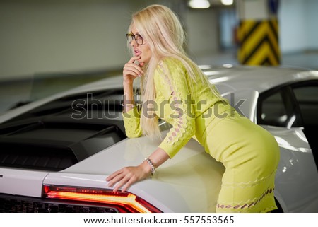 Slim blonde woman in yellow dress stands leaning on trunk of modern white car at underground parking.