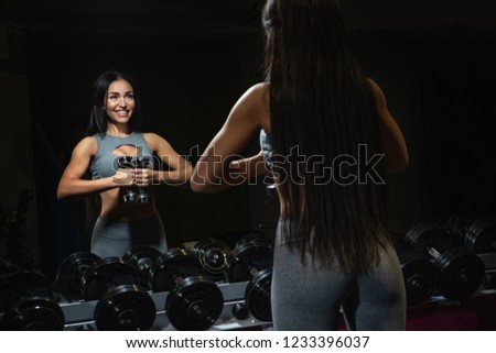 Slim bodybuilder girl lifts heavy dumbbell standing in front of the mirror while training in the gym.