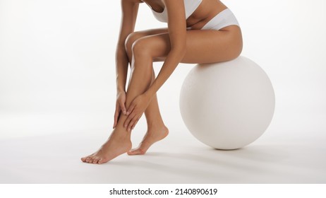 Slim female model in white underwear strokes her leg sitting on big white ball on white background | Smooth legs and unwanted hair removal commercial concept - Shutterstock ID 2140890619