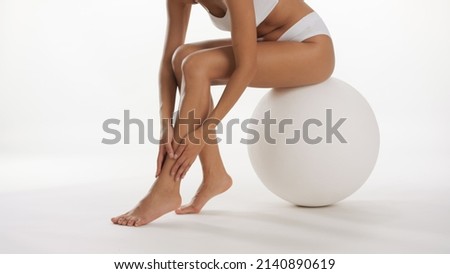 Slim female model in white underwear strokes her leg sitting on big white ball on white background | Smooth legs and unwanted hair removal commercial concept