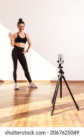 Slim Female Recording Tutorial Video For Fitness Vlog, Waving Hand, Saying Hi To Followers, Wearing Black Sports Top And Tights. Full Length Studio Shot Illuminated By Sunlight From Window.