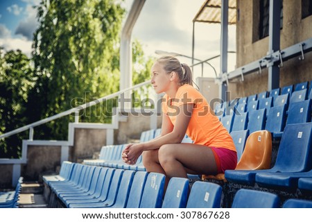 Slim female in sportswear sitting on plastic blue seat on the stadium.