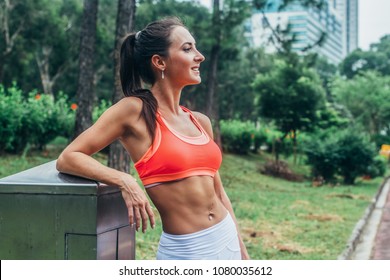 Slim Fitness Brunette Woman With Six Pack Abs Wearing Pink Sport Bra Standing In City Park Relaxing After Workout Looking Away From The Camera