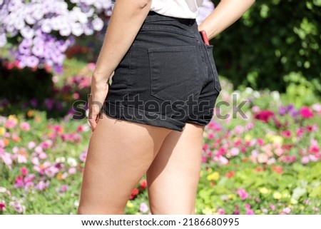 Slim girl in black jeans shorts standing on city street on flowers background. Female fashion in hot summer weather