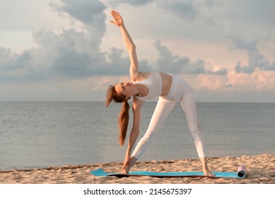 Slim girl doing yoga at sunset on sandy shore. Practice yoga on the beach on sea background. Triangle pose, trikonasana.