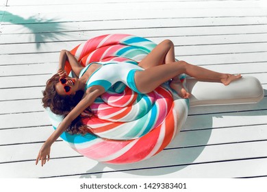 Slim girl having fun on colorful Inflatable lollipop pool float. Bikini model in bright swimwear sun tanning on tropical bech at summer day. 