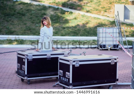 slim girl in a white coat  standing next to a flightcase with equipment. Preparing the stage for a concert in the open air.