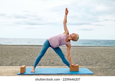 Slim Good Looking Woman Practicing Yoga At The Beach Using Wooden Yoga Blocks For More Comfortable And Easier For Practise While Doing Exercise.