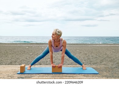 Slim Good Looking Woman Practicing Yoga At The Beach Using Wooden Yoga Blocks For More Comfortable And Easier For Practise While Doing Exercise.