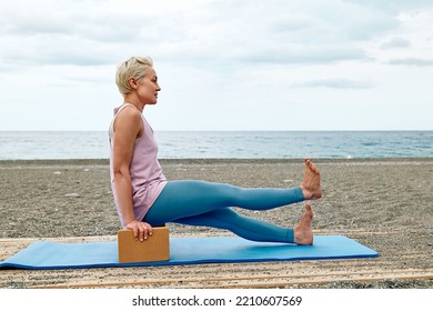 Slim Good Looking Woman Practicing Yoga At The Beach Using Wooden Yoga Blocks For More Comfortable And Easier For Practise While Doing Exercise.