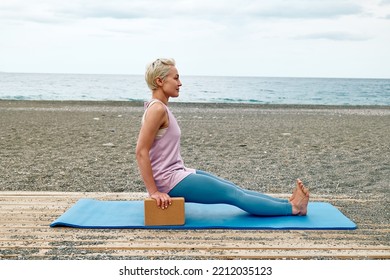 Slim Good Looking Woman Practicing Yoga At The Beach Using Wooden Yoga Blocks For More Comfortable And Easier For Practise While Doing Exercise.