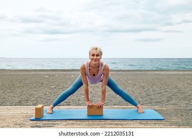 Slim Good Looking Woman Practicing Yoga At The Beach Using Wooden Yoga Blocks For More Comfortable And Easier For Practise While Doing Exercise.