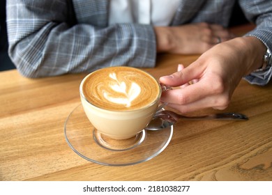 A slim hand holds a cup of coffee capuchino with a beautiful image on the top on the wooden table close up