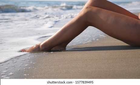 Slim Legs Of Woman Lying On Tropical Beach With Splashing Waves. Ocean Tides Washing Over Tanned Female Feet. Young Girl Relaxing On Coast During Summer Vacation. Summer Holiday Concept. Slow Mo