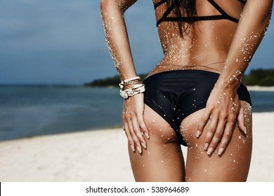 Slim luxury girl in a black bikini on the beach. Photos from behind. Perfect tanned body, sexy ass, perfect figure. Rest on a tropical island. Golden bracelets Photo in low key. Vogue Style