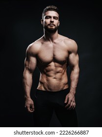 Slim muscular male model at black background. Fitness shirtless guy in black sport pants posing in studio. Man with six pack abs looking at camera.