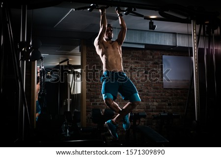 Slim muscular man doing pullups shirtless in the gym and listening to music on the headphones, high contrast image