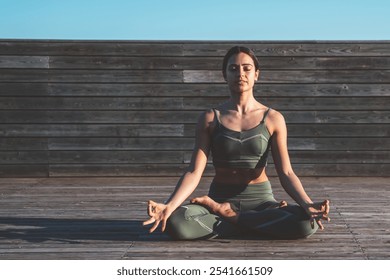 Slim peaceful ethnic female yogi meditating while sitting in lotus asana with closed eyes on wooden terrace in sunny morning time - Powered by Shutterstock