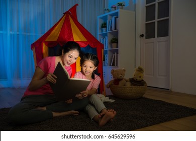 Slim Smiling Lady And Happy Leisurely Child Sitting In Front Of Toy Tent Reading Story Comic Book Together On Bed Room At Night.