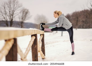 Slim Sportswoman Leaning On Fence In Nature At Snowy Winter Day And Doing Stretching Exercises. Warmup Exercise, Outdoor Fitness, Winter Fitness